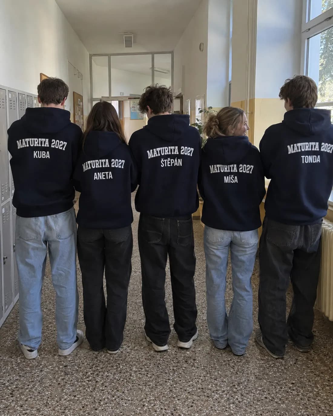 Maturanti zezadu v tmavých mikinách, na zádech nápis Maturita 2027 a jména